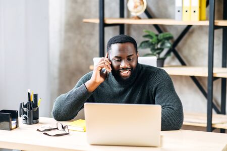 Cheerful African-American office employee sits at the desk with a laptop in modern office and talks on the phoneの写真素材