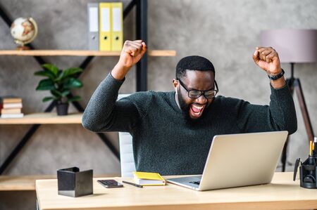 Success at work, good deal. African-American guy looks at the laptop screen, screams excitedly and raises his fists in a victory gesture