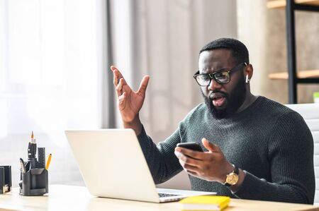 Confused African-American guy looks at phone screen while sitting at the office desk with a laptop, he read some bad news or messageの写真素材