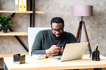 An african-american guy in smart casual wear and glasses is typing messages on smartphone while working with a laptop in contemporary officeの写真素材