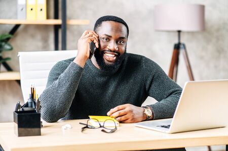 Smiling African-American guy in glasses sits at the desk in modern office, he use trendy laptop for a work and is talking on the smartphone. Front viewの写真素材