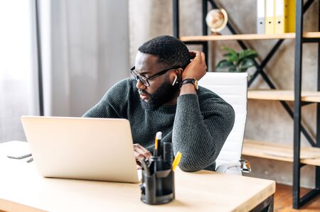 Pensive african-american office employee at the desk is watching focused at the laptop screenの写真素材