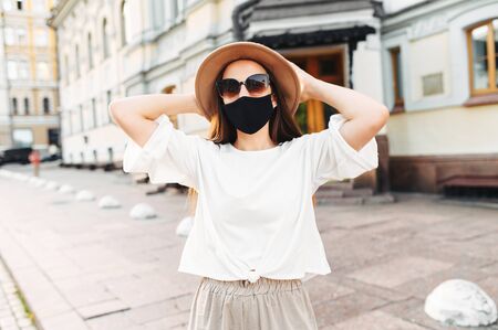 Young woman in a stylish hat and sunglasses posing with a black medical mask on her face outdoorsの写真素材