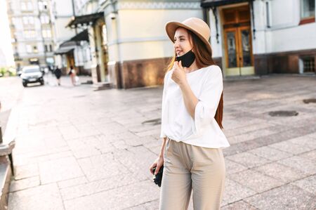 Tourist in the city after quarantine. Young attractive girl sightseeing with lowered protective mask on the chin, she stands in the downtown with a camera in hands in the city centerの写真素材
