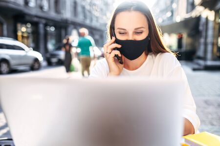 Epidemia in the city. Young woman freelancer with a protective medical mask on the face is working with laptop on the outdoor summer terrace cafe, she sits at the table and talking on the phoneの写真素材