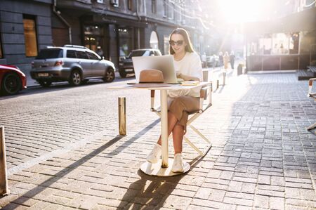 Young attractive woman sits at the table of outdoor cafe on a cozy european street with a laptop in warm natural sunlightの写真素材