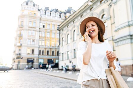 An attractive young girl is walking along a city street, talking on the phone and laughing. Close portrait of a stylish happy young woman in a hat and with a paper shopping bagの写真素材