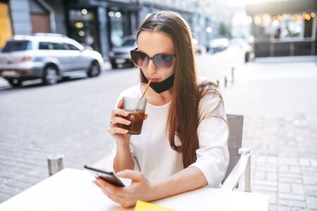 Attractive girl drinks cold coffee in a summer cafe after quarantine. Young woman in sunglasses lowered a medical mask on her chin, sits and chill in a summer cafe with a smartphone in handsの写真素材