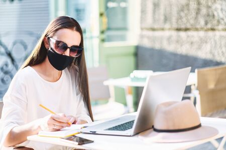 Female student is watching lectures on laptop and taking notes while sitting at the table with a medical mask on the face in the summer street cafeの写真素材