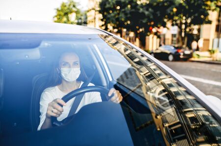 View through the windshield of a woman in a medical mask driving a car. Young focused woman holds hands on the steering wheelの写真素材