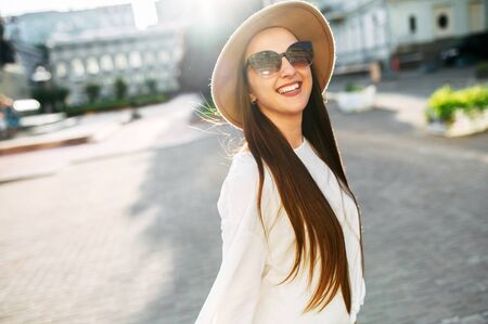 Cheerful beautiful brunette in a stylish hat and sunglasses looks at the camera and laughs happily. Stylish young woman with long hair walks around the cityの写真素材
