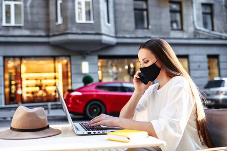 Epidemia in the city. Young woman freelancer with a protective medical mask on the face is working with laptop on the outdoor summer terrace cafe, she sits at the table and talking on the phoneの写真素材