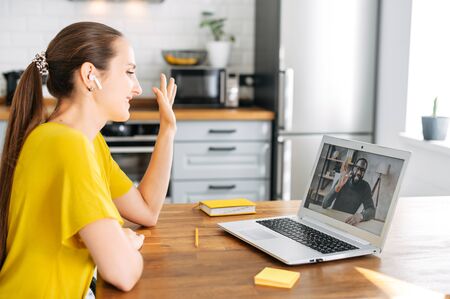 A young woman is working from home and talking online with a coworkers, employees. Woman waving to multiracial people on the laptop screen. Video meetingの写真素材