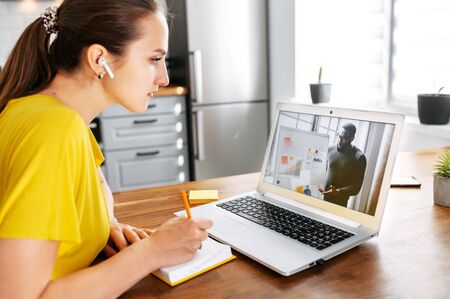 An African-American male teacher conducts webinar, online classes on the laptop screen indoor, young woman is watching and taking notes in the kitchen at home. Online studying conceptの写真素材