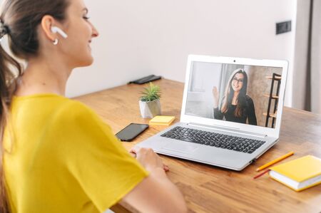 A young woman sits indoor and using app on laptop for video call to female coworker, colleague or friend. Two young women in casual wear talking online via video connectionの写真素材