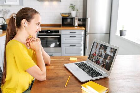 Remote work, virtual meeting. Young woman with laptop in the kitchen is talking with coworkers, employees from homeの写真素材