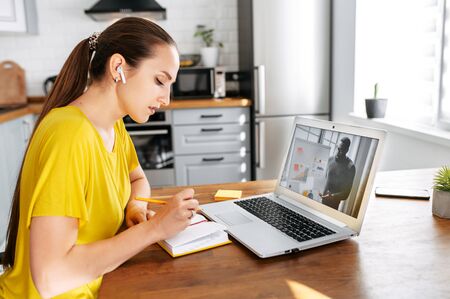 An African-American male teacher conducts webinar, online classes on the laptop screen indoor, young woman is watching and taking notes in the kitchen at home. Online studying conceptの写真素材
