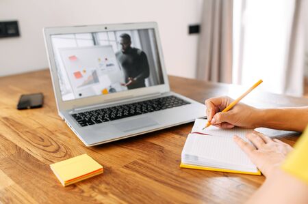 An African-American man conducts webinar, online classes on the laptop screen indoor, female hands are taking notes in notebook at the table in the kitchen at home. Online studying conceptの写真素材