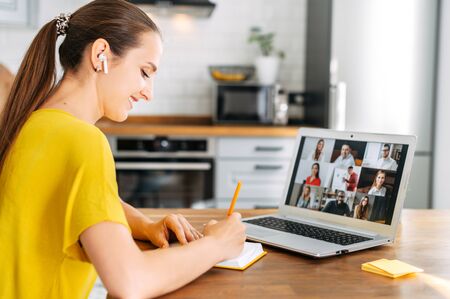 Side view of young woman watching on laptop display with a group of multiracial people on it and writing notes. Video call, online conference, webinarの写真素材
