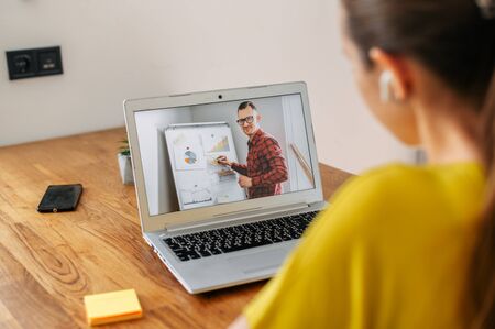 Webinars, online learning, online classes. A female student using laptop for watching lectures online, a woman sits at the table and writing in notebook, a male teacher with flip chart on the screenの写真素材