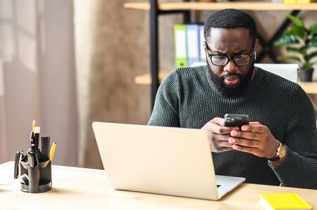 Puzzled black guy with glasses is typing a message on the phone, he is sitting in front of a laptop in the officeの写真素材