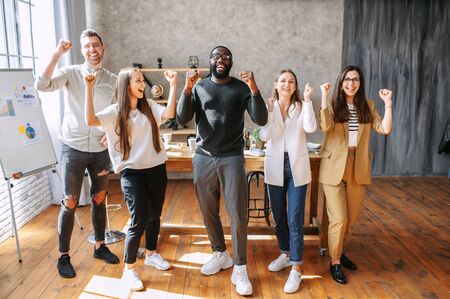 Team of young successful multiracial colleagues are happy, they stand in the office laughing with hands up. Long shot, casual officeの写真素材