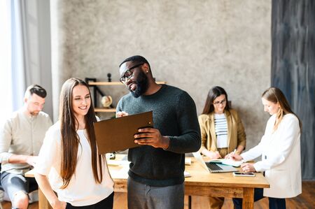 A working process of two colleagues working in office. Smiling young woman stands with an afro-american guy, they studying something on a pad. Multiracial team in office on the backgroundの写真素材
