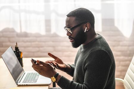 Confused African-American guy looks at phone screen while sitting at the office desk with a laptop, he read some bad news or messageの写真素材
