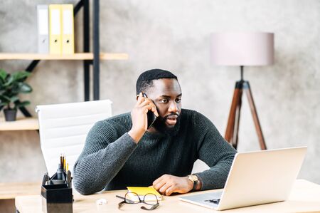 Young black man is solving business tasks on the phone, concentrated guy sits at the desk with laptop in contemporary office and talking on the phoneの写真素材