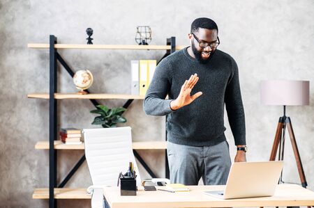 Young black employee is using video connection for virtual meeting with co-workers, a guy standa in front of laptop in contemporary office, greeting and waving into webcamの写真素材