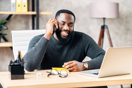 Cheerful African-American office employee sits at the desk with a laptop in modern office and talks on the phoneの写真素材