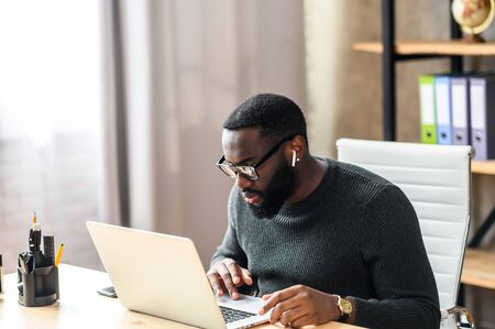 Concentrated black guy in smart casual wear using laptop for work or studying. African-American guy in glasses focused looks at laptop screenの写真素材