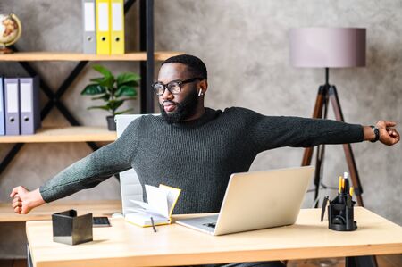 Smart African-American guy with glasses tired of working with a laptop in the office, he sits in a chair and stretchesの写真素材