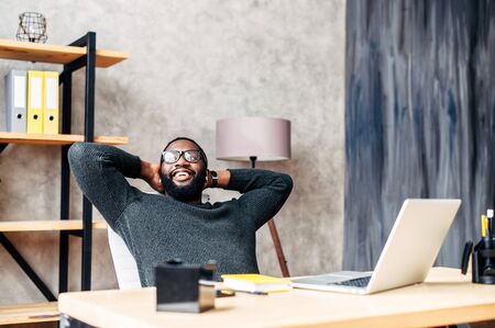 Take a break on a workplace. A young African-American guy leaned back in his chair relaxed, he is pleased with the work doneの写真素材