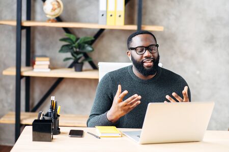 Video call. Attractive African-American guy is using laptop for video communication, a guy in glasses sits at the table in modern office, looking into webcam and talking onlineの写真素材