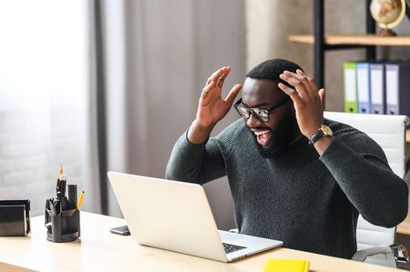 Overjoyed African man raised hands up, a guy in glasses excited and happy with a good news or deal, he looks at laptop screen with triumphの写真素材