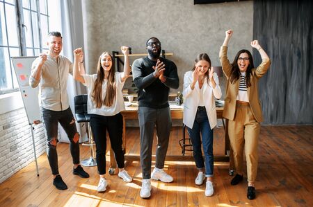 Long shot of young successful multiracial team with their hands up in cosy office. They are laughing and have happy look on their facesの写真素材