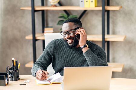 Smiling African-American guy in glasses sits at the desk in modern office and use trendy laptop for a work. A young man is talking on the smartphone and writting. Front viewの写真素材