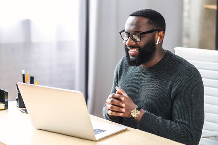 Side view of African-American office employee talking via video connection while sitting at the desk. A guy using laptop for video call in contemporary office spaceの写真素材