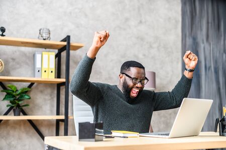 Success at work, good deal. African-American guy looks at the laptop screen, screams excitedly and raises his fists in a victory gestureの写真素材