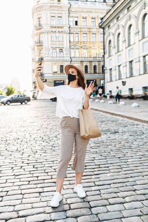 A stylish girl in mask is taking selfie on the phone or talking via video call outdoors. Young woman in mask is waving into smartphone. Full length, vertical shotの写真素材