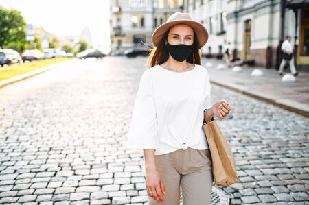 A young woman in a protective mask walks along a city street with a paper shopping bag in her hands. City walking and shopping after quarantineの写真素材