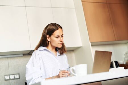 Concentrated young woman freelancer is working with a laptop from home at a table in the kitchen. Remote Work Conceptの写真素材