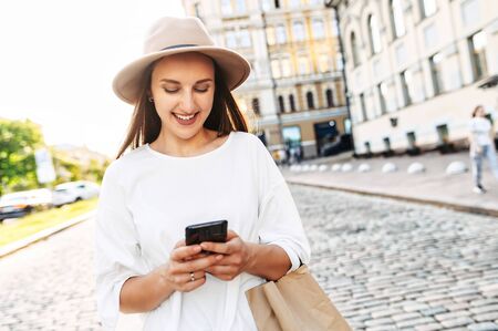 Attractive young girl with a smartphone in her hands on a city street, chatting and laughing. Close portrait of stylish happy young woman in hat and with paper shopping bag typing message on phoneの写真素材
