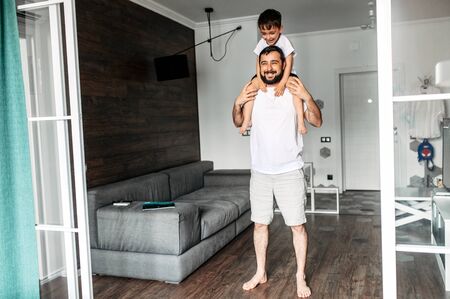 Young father his little son spend time together at home. Dad holds toddler on the shoulders in cozy living room. Happy togetherの写真素材