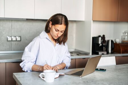 Concentrated young woman freelancer is working with a laptop from home at a table in the kitchen. Remote Work Conceptの写真素材
