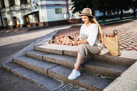 Attractive young girl with a smartphone in her hands sits on a stairs in the city, chatting on the phone and laughing. Stylish happy young woman in hat and with paper shopping bag typing messageの写真素材