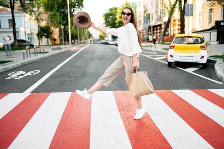 Funny shot of cheerful young woman is walking on zebra crosswalk. A stylish happy girl waving hat and looking into camera with a smileの写真素材