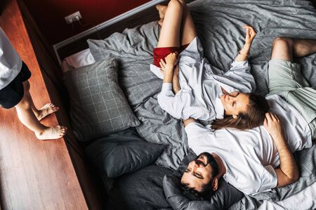 Young family spends time together at home. Mother and father lie in the bedroom on the bed and laugh, they are looking at son with love. The kid runs on the bed, his face is not visibleの写真素材