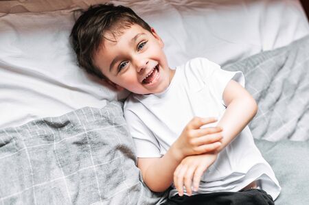 Happy childhood. Cute toddler boy lying on the bed, laughing and looking at the cameraの写真素材